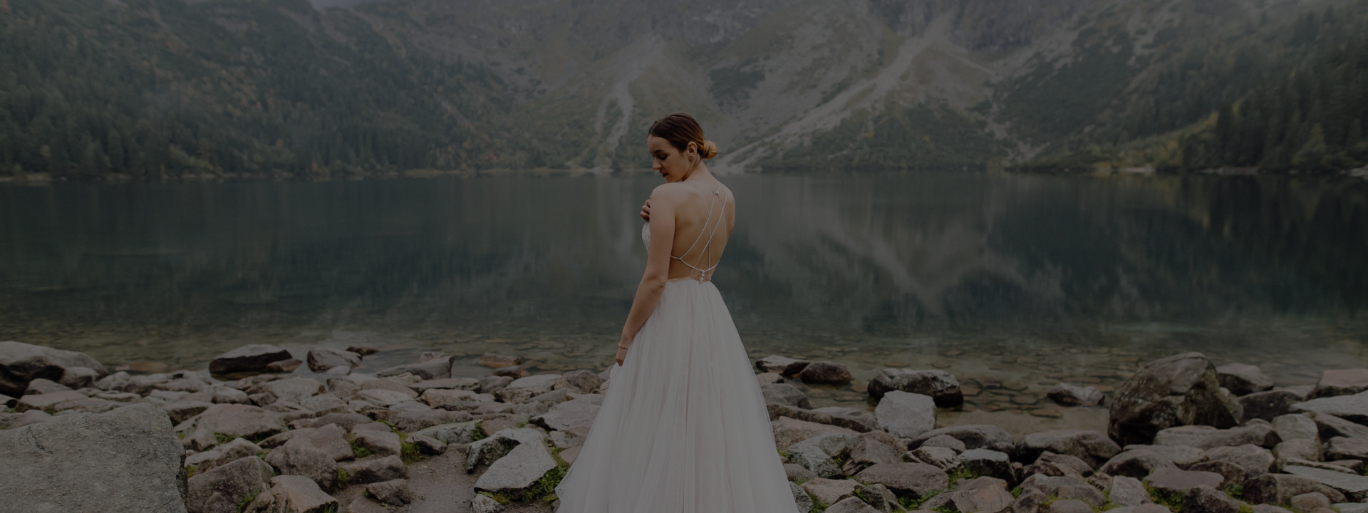 A woman in a white dress standing by a lake with mountains in the background